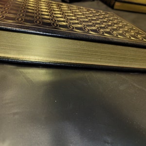 May include: A black leather-bound book with gold-colored edges and a decorative pattern on the cover. The book is lying on a dark gray surface.