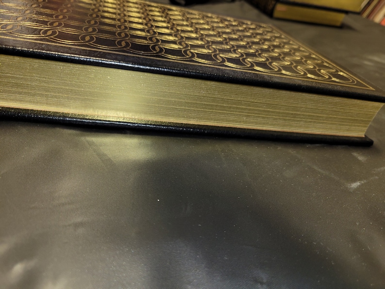 May include: A black leather-bound book with gold-colored edges and a decorative pattern on the cover. The book is lying on a dark gray surface.