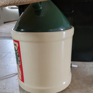 May include: A white ceramic crock with a green lid and a red and green label that says "Springing" with a picture of a frog. The crock has a white cord coming out of the bottom.
