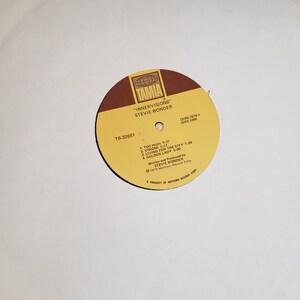 Puede incluir: Una etiqueta de disco amarilla y marr&oacute;n con el texto "TAMLA" en un c&iacute;rculo blanco. El texto "INNERVISIONS" de STEVIE WONDER" est&aacute; impreso debajo del c&iacute;rculo. El n&uacute;mero de disco T6-32651 y la lista de pistas del lado uno tambi&eacute;n est&aacute;n impresos en la etiqueta.