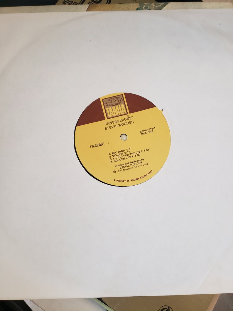 Puede incluir: Una etiqueta de disco amarilla y marr&oacute;n con el texto "TAMLA" en un c&iacute;rculo blanco. El texto "INNERVISIONS" de STEVIE WONDER" est&aacute; impreso debajo del c&iacute;rculo. El n&uacute;mero de disco T6-32651 y la lista de pistas del lado uno tambi&eacute;n est&aacute;n impresos en la etiqueta.