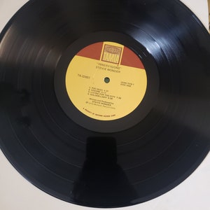 Puede incluir: Un disco de vinilo negro con una etiqueta amarilla. La etiqueta tiene el texto "TAMLA", "INNERVISIONS", "STEVIE WONDER", "CSAS-7576-1", "SIDE ONE", "T6-32651", "1. TOO HIGH 4:37", "2. VISIONS 5:17", "3. LIVING FOR THE CITY 7:26", "4. GOLDEN LADY 5:00", "Written and Produced by STEVIE WONDER", "1972 Motown Record Corp.", y "A PRODUCT OF MOTOWN RECORD CORP.".