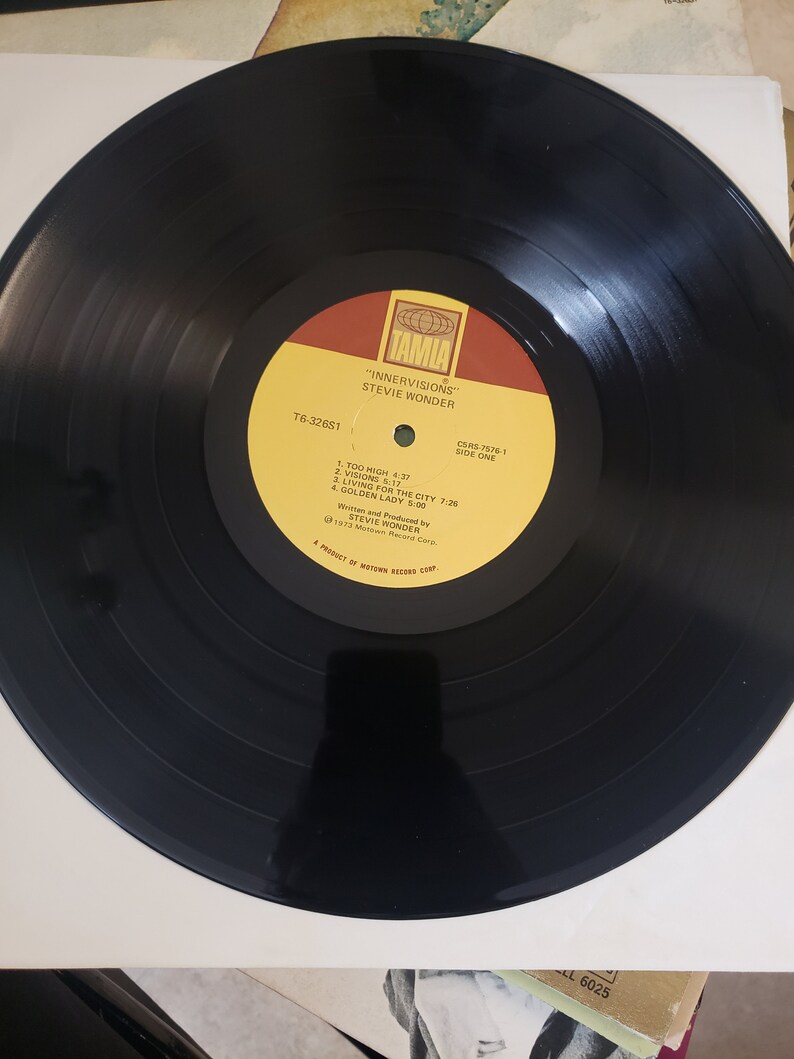Puede incluir: Un disco de vinilo negro con una etiqueta amarilla. La etiqueta tiene el texto "TAMLA", "INNERVISIONS", "STEVIE WONDER", "CSAS-7576-1", "SIDE ONE", "T6-32651", "1. TOO HIGH 4:37", "2. VISIONS 5:17", "3. LIVING FOR THE CITY 7:26", "4. GOLDEN LADY 5:00", "Written and Produced by STEVIE WONDER", "1972 Motown Record Corp.", y "A PRODUCT OF MOTOWN RECORD CORP.".