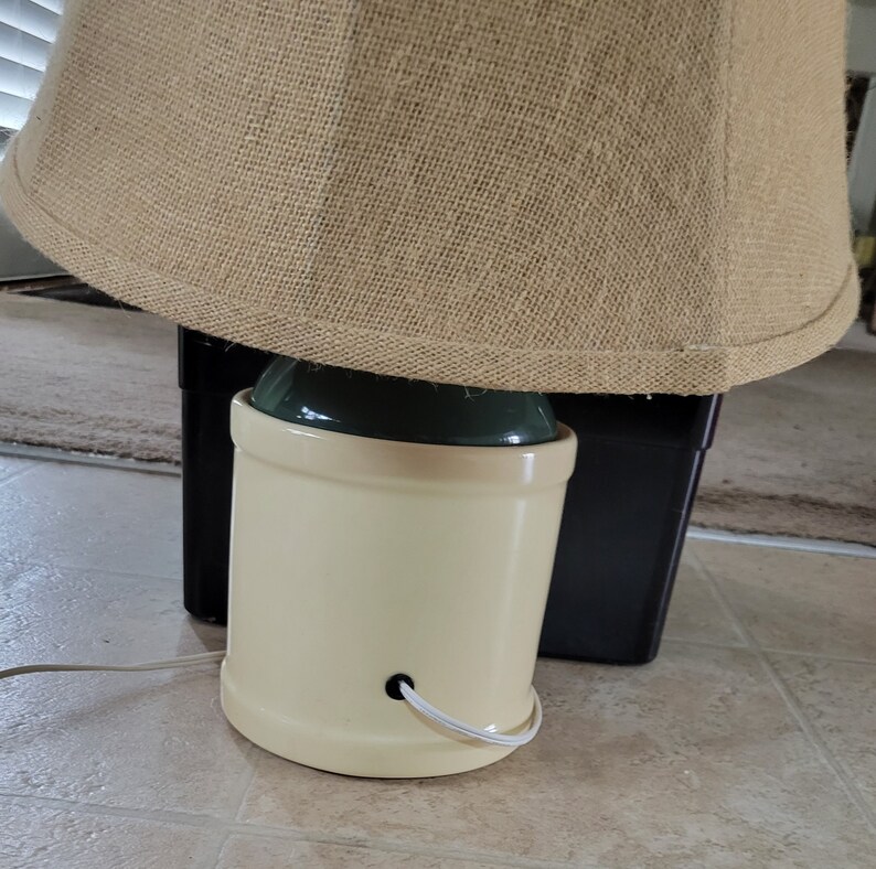 May include: A beige ceramic lamp base with a green top and a burlap lampshade. The lamp is plugged in and ready to use.