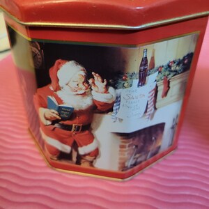Puede incluir: Lata octogonal roja y dorada con una escena navide&ntilde;a que representa a Pap&aacute; Noel leyendo un libro, una chimenea y una botella de Coca-Cola. El texto de la carta dice "Querido Pap&aacute; Noel, por favor, tr&aacute;enos m&aacute;s Coca-Cola".