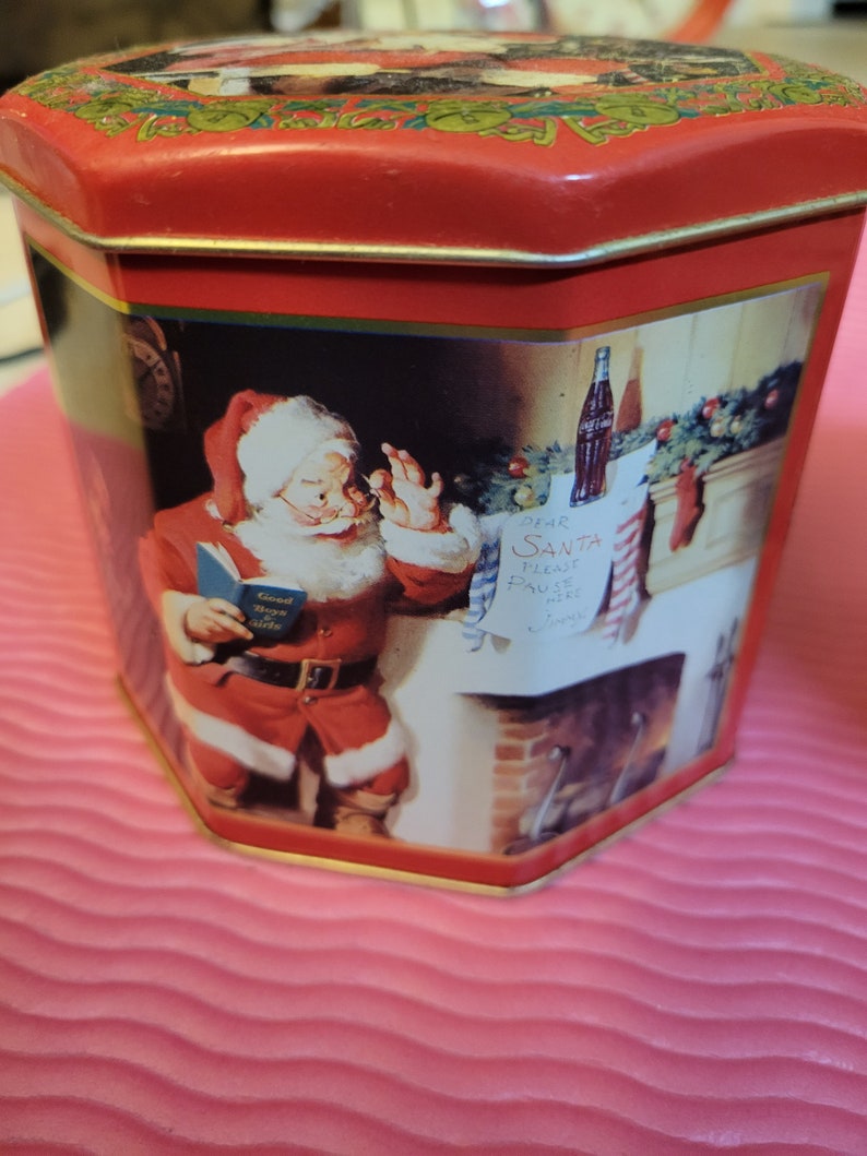 Puede incluir: Lata octogonal roja y dorada con una escena navide&ntilde;a que representa a Pap&aacute; Noel leyendo un libro, una chimenea y una botella de Coca-Cola. El texto de la carta dice "Querido Pap&aacute; Noel, por favor, tr&aacute;enos m&aacute;s Coca-Cola".