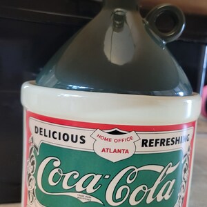 May include: A green and white ceramic Coca-Cola dispenser with a red, white, and green label. The label reads "Delicious, Home Office Atlanta, Refreshing, Coca-Cola, Trade Mark Registered, To Dispense, Draw one ounce of Coca-Cola in seven ounce glass, then fill glass with large stream of soda water stirring with a spoon that Coca-Cola may be thoroughly mixed. The Coca-Cola Company All Rights Reserved. The Coca-Cola Co. Atlanta."