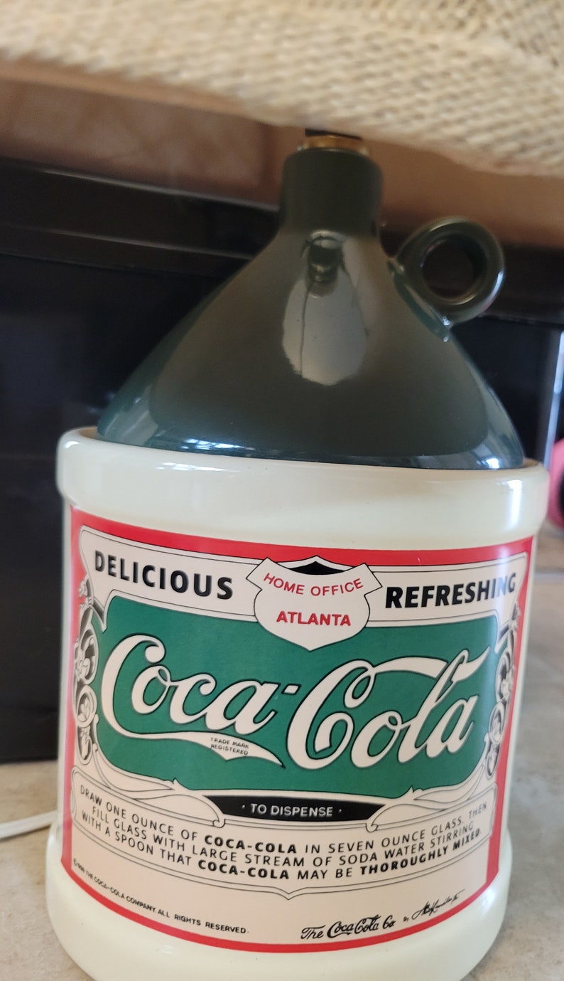 May include: A green and white ceramic Coca-Cola dispenser with a red, white, and green label. The label reads "Delicious, Home Office Atlanta, Refreshing, Coca-Cola, Trade Mark Registered, To Dispense, Draw one ounce of Coca-Cola in seven ounce glass, then fill glass with large stream of soda water stirring with a spoon that Coca-Cola may be thoroughly mixed. The Coca-Cola Company All Rights Reserved. The Coca-Cola Co. Atlanta."