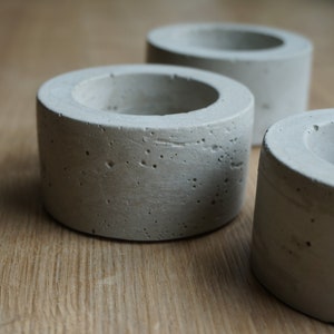 Könnte beinhalten: Drei graue Beton-Kerzenhalter mit einer rauen, strukturierten Oberfläche. Die Halter sind zylindrisch geformt und haben eine kleine, runde Öffnung oben.