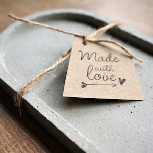 Könnte beinhalten: Eine graue, ovale Betonablage mit rauer Textur. Ein braunes Papieretikett mit Schnur gebunden, das "Made with love" mit einem Pfeil und einem Herzsymbol trägt. Die Ablage ist ein dekorativer Artikel.