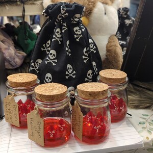May include: Four small glass jars filled with red liquid and red dice. Each jar has a cork lid and a brown paper tag that reads "Potion of Healing". A black fabric bag with a skull and crossbones pattern is in the background.