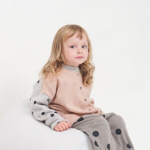 Puede incluir: Una niña pequeña sentada en un puf blanco, lleva un suéter marrón claro con mangas de lunares negros y pantalones a juego.