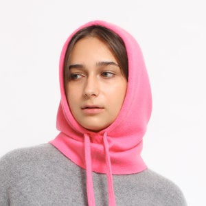 May include: A pink fleece hooded neck warmer with a drawstring. The neck warmer is worn by a person with brown hair.