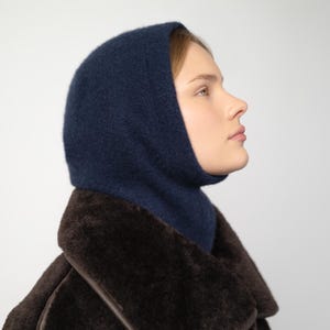 Puede incluir: Una capucha de punto azul marino y un abrigo marrón oscuro. La capucha cubre la cabeza y el cuello, con una abertura para la cara. El abrigo tiene una textura similar a la piel marrón oscuro. El fondo es blanco liso.