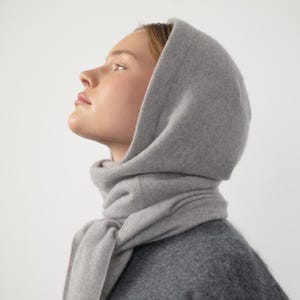 Può includere: Un set di sciarpa e cappuccio grigio chiaro. Il cappuccio è drappeggiato sulla testa e la sciarpa è avvolta attorno al collo. Il tessuto sembra morbido e caldo, con una trama sottile. Lo stile generale è semplice ed elegante.