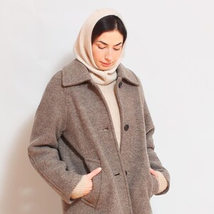 May include: A brown wool coat with a large collar and a button closure. The coat is worn over a white turtleneck sweater and a cream-colored hooded scarf.