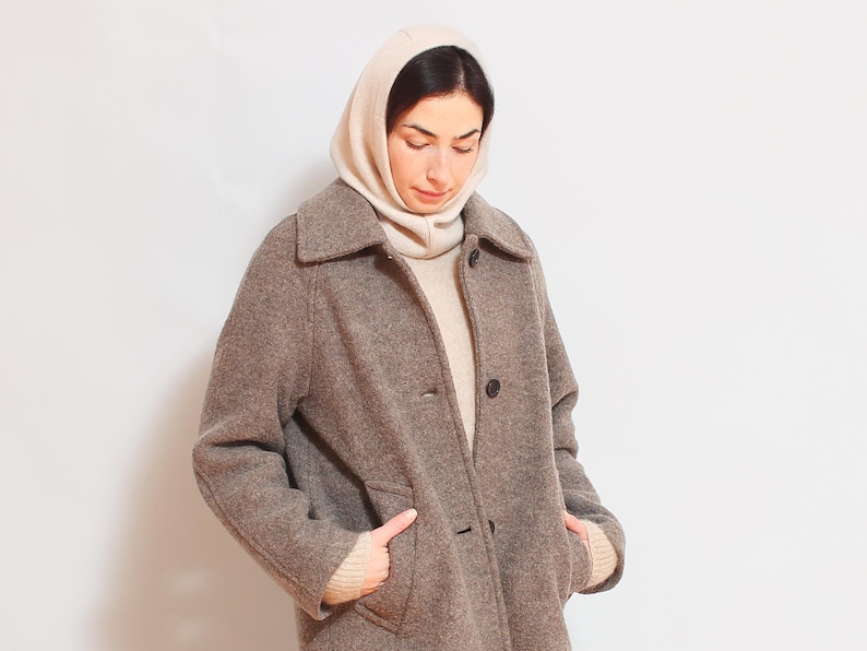 May include: A brown wool coat with a large collar and a button closure. The coat is worn over a white turtleneck sweater and a cream-colored hooded scarf.