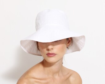 May include: A white sun hat with a wide brim. The hat is made of a soft, textured fabric and has a rounded crown. The brim is slightly wavy. The hat is worn by a person with fair skin and light-colored hair.