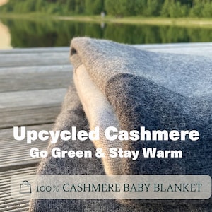 Può includere: Una copertina per neonati in cashmere grigio piegata su un molo di legno. Il testo "Upcycled Cashmere Go Green & Stay Warm 100% CASHMERE BABY BLANKET" è visibile sulla copertina.