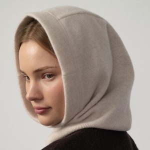 May include: A light beige hooded scarf, worn over a dark brown garment. The hood is made of a soft, textured material and frames the face. The scarf appears to be a warm, winter accessory.