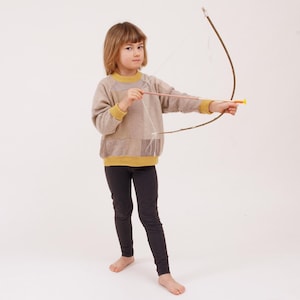 Puede incluir: Una persona joven con un suéter marrón y amarillo y leggings negros sostiene un arco y una flecha marrones. El arco está hecho de madera y tiene una cuerda atada. La flecha está hecha de madera y tiene una punta amarilla.
