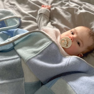 Peut inclure: Un bébé portant une couverture à motifs bleu clair et blanc avec une sucette dans la bouche. Le bébé est allongé sur une couverture grise.