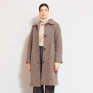 May include: A brown wool coat with a button closure and a white turtleneck sweater underneath. The person is wearing a white knitted hat.