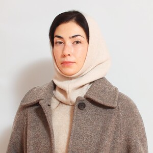 May include: A woman wearing a beige knitted turtleneck sweater and a brown wool coat. The sweater has a high neck and covers the woman's head.