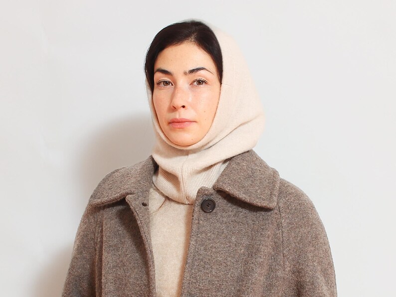May include: A woman wearing a beige knitted turtleneck sweater and a brown wool coat. The sweater has a high neck and covers the woman's head.