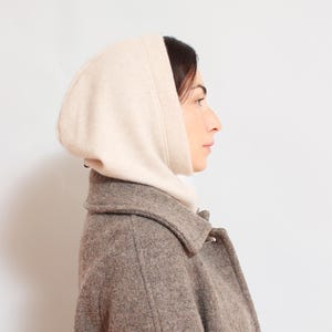 May include: A woman wearing a beige hooded sweater over a brown wool coat.