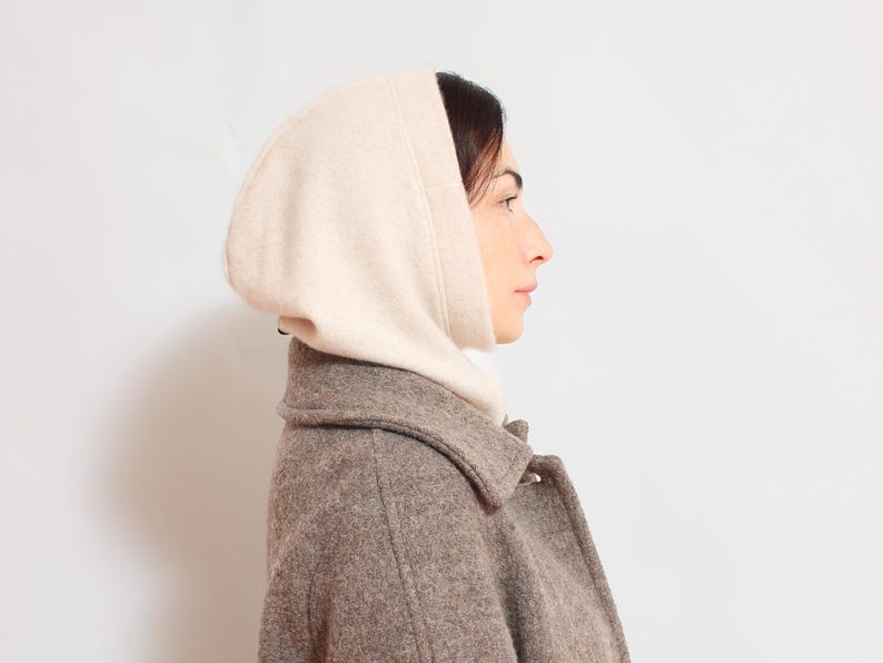 May include: A woman wearing a beige hooded sweater over a brown wool coat.
