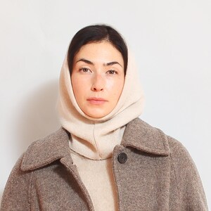 May include: A woman wearing a beige knitted turtleneck sweater and a brown wool coat. She is wearing a beige knitted cowl neck scarf.