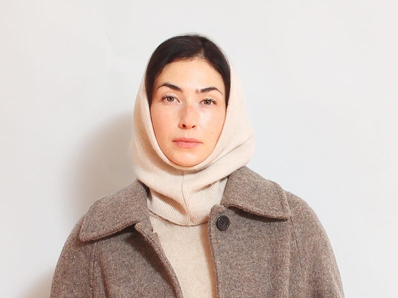 May include: A woman wearing a beige knitted turtleneck sweater and a brown wool coat. She is wearing a beige knitted cowl neck scarf.
