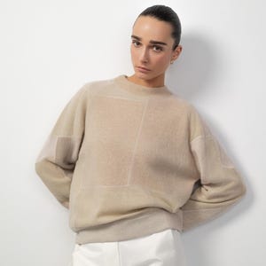 Peut inclure: Un pull beige avec un motif géométrique. Le pull a un col rond et des manches longues. Le modèle porte un pantalon blanc. Le pull est fait d'une matière douce et texturée.