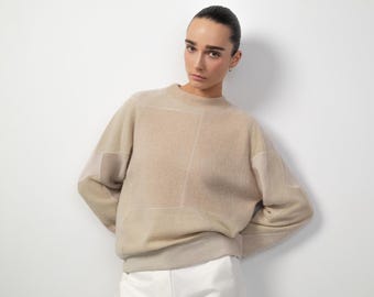 Könnte beinhalten: Ein beiger Pullover mit geometrischem Muster. Der Pullover hat einen Rundhalsausschnitt und lange Ärmel. Das Model trägt eine weiße Hose. Der Pullover ist aus einem weichen, strukturierten Material gefertigt.