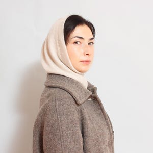 May include: A woman wearing a brown wool coat with a white knitted scarf. The scarf is tied around her neck and covers her head.