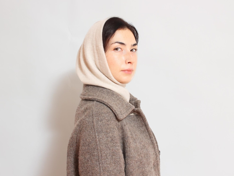 May include: A woman wearing a brown wool coat with a white knitted scarf. The scarf is tied around her neck and covers her head.