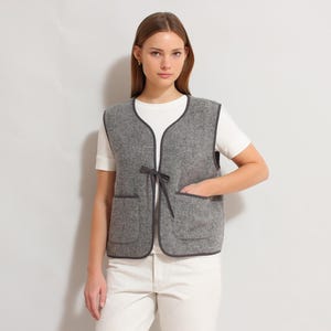Op de afbeelding: Een grijze vest met een donkere bies en twee voorzakken. De vest heeft een striksluiting en wordt over een wit t-shirt gedragen. De vest is gemaakt van een textuurstof.