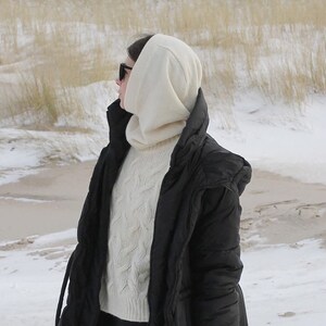 May include: A woman wearing a black puffer jacket with a white hooded sweater underneath. She is wearing sunglasses and standing in a snowy landscape.