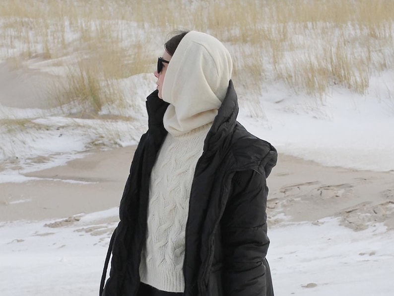May include: A woman wearing a black puffer jacket with a white hooded sweater underneath. She is wearing sunglasses and standing in a snowy landscape.