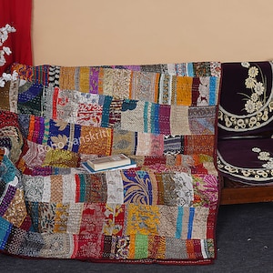 May include: A colorful patchwork quilt made from recycled saris, featuring a variety of patterns and colors. The quilt is draped over a wooden chair with a purple cushion. The quilt is approximately 60 inches by 80 inches.