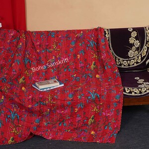 May include: Red kantha quilt with a floral and bird pattern. The quilt is folded and draped over a wooden chair with a purple cushion. The text "Boho Sanskriti" is visible on the quilt.