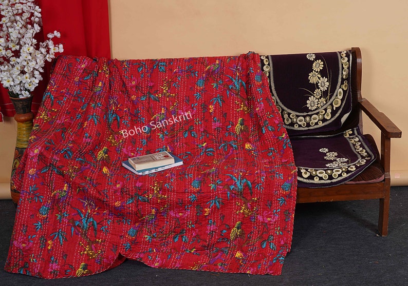 May include: Red kantha quilt with a floral and bird pattern. The quilt is folded and draped over a wooden chair with a purple cushion. The text "Boho Sanskriti" is visible on the quilt.