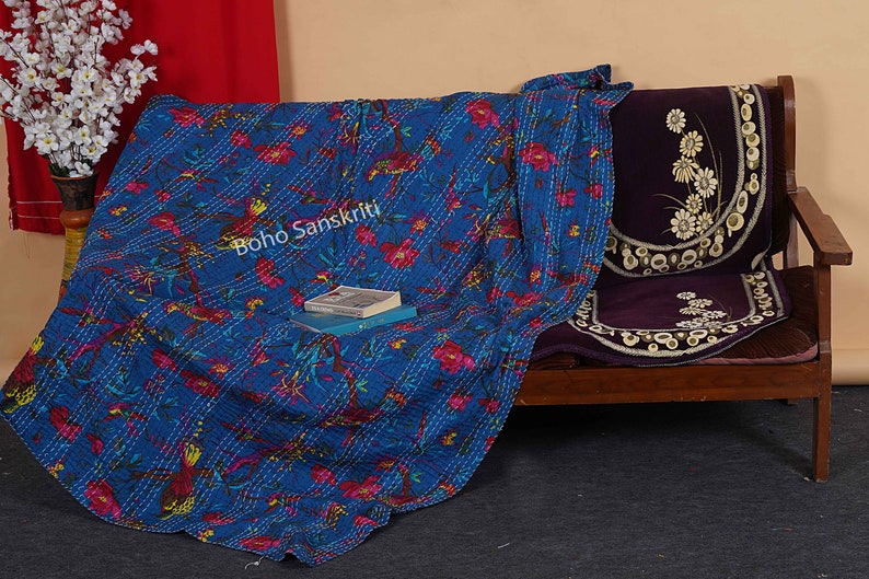 May include: A blue kantha quilt with a floral pattern and white stitching. The quilt is draped over a wooden bench with a purple cushion. The text "boho sanskriti" is visible on the quilt.