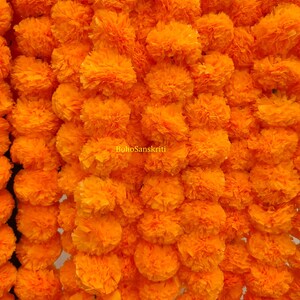 Op de afbeelding: Een close-up van talrijke feloranje goudsbloemen, dicht op elkaar gepakt. De afbeelding benadrukt de textuur en kleur van de bloemen, waarbij elke bloem meerdere lagen bloemblaadjes laat zien. De tekst "BohoSanskriti" is zichtbaar.