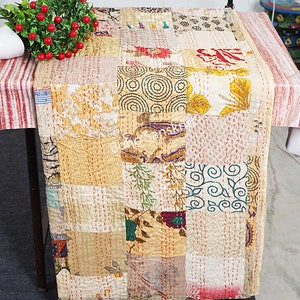 May include: A beige patchwork table runner with a variety of floral and geometric patterns. The runner is made of kantha fabric, a traditional Indian hand-stitched fabric.