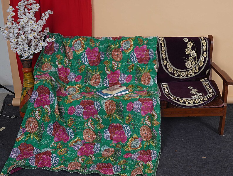 May include: A green kantha quilt with a floral and pineapple pattern. The quilt is draped over a wooden bench with a purple cushion cover.