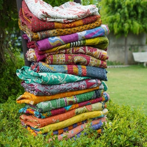 Pode incluir: Uma pilha de colchas kantha coloridas com padrões florais e geométricos. As colchas estão dobradas e empilhadas em uma cadeira de madeira na frente de um arbusto verde. O texto "boho sanskriti" é visível em duas das colchas.