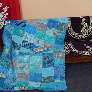 May include: A patchwork quilt made of various shades of blue fabric. The quilt is folded and placed on a wooden bench with a purple cushion. A book is placed on top of the quilt.