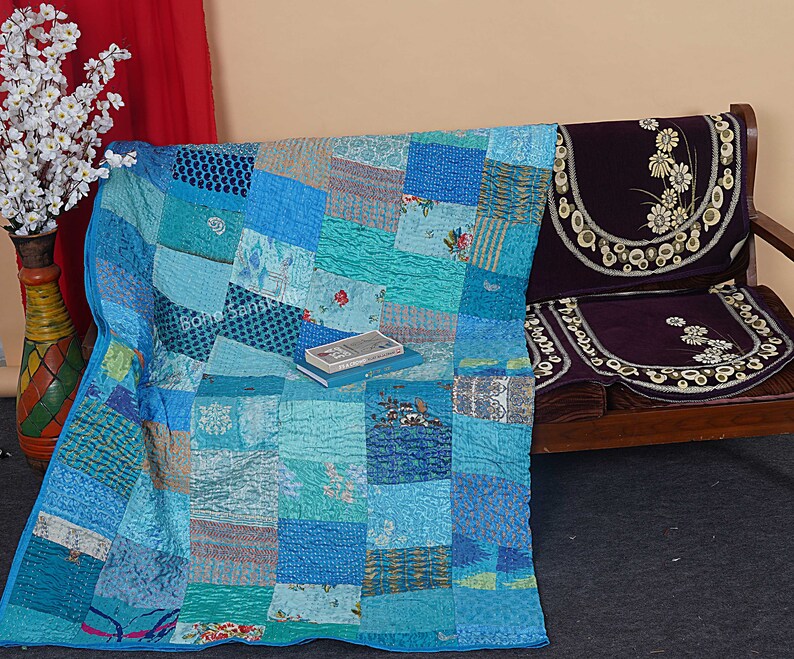 May include: A patchwork quilt made of various shades of blue fabric. The quilt is folded and placed on a wooden bench with a purple cushion. A book is placed on top of the quilt.